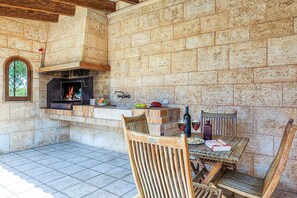 An outdoor patio with a stone wall, a brick fireplace, a dining table with chairs, and a window with a decorative iron grille.