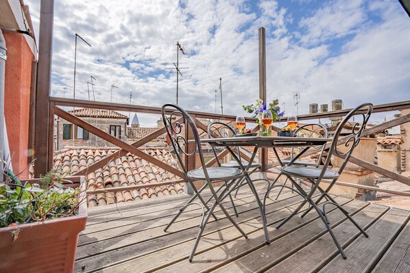 A rooftop terrace with a round table and four chairs, a potted plant, and a table setting with a glass of wine and a vase of flowers.