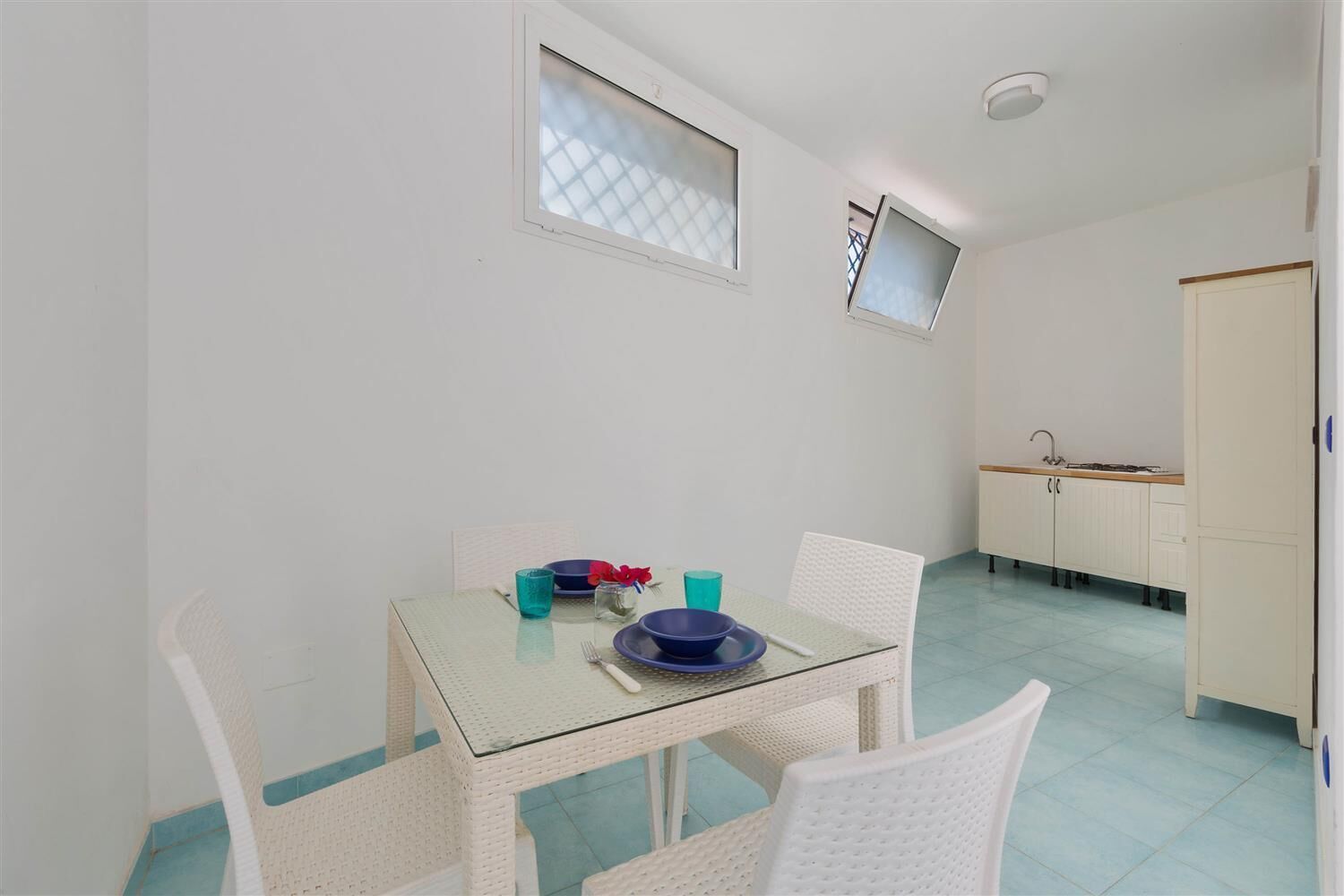 Un comedor con una mesa de vidrio, sillas de mimbre blanco y una cocina al fondo.
