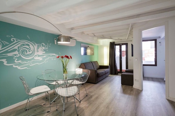 A modern living room with a glass dining table, a sofa, and a mural on the wall.