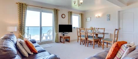 Un salon comprenant un ensemble de canapés, un téléviseur, une table à manger avec des chaises, et une vue sur la mer grâce à de grandes fenêtres.