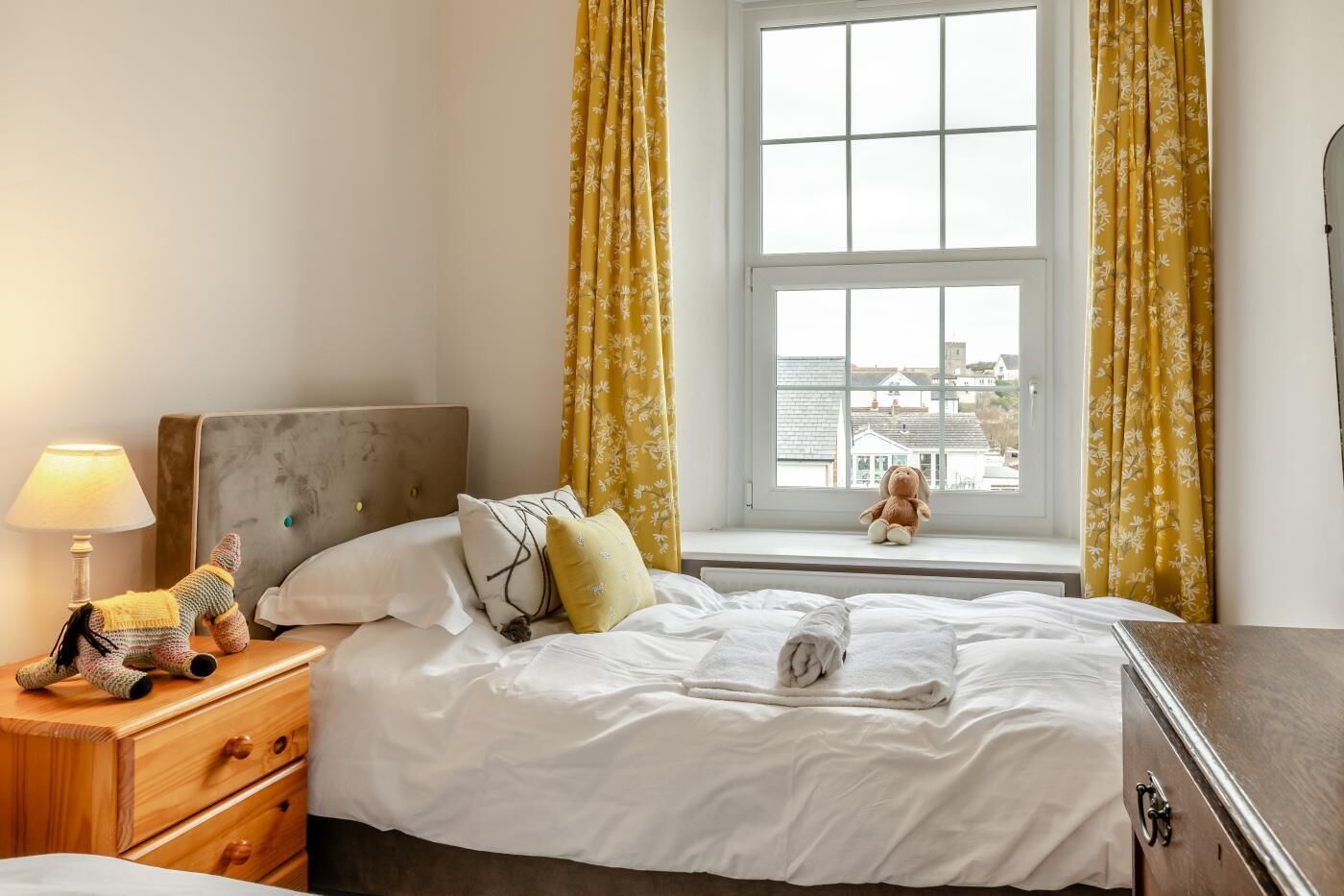 En pænt redt seng med hvide sengetøj, et trænatbord med en lampe, en kommode og et vindue med gule gardiner.