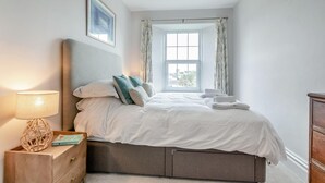 A bedroom with a bed, a bedside lamp, a nightstand, a window with curtains, and a framed picture on the wall. - 2 Bed in Mortehoe (oc-mail1) (Mortehoe)
