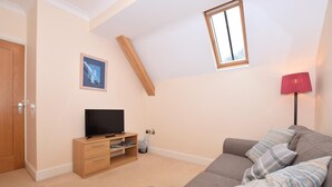 Ein Zimmer mit Fernseher auf einem Holzboden, ein graues Sofa mit gemusterten Kissen, eine rote Stehlampe und ein Fenster mit Holzrahmen.
