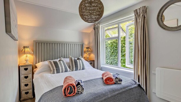 A neatly made bed with striped pillows, a round woven light fixture, and a window with curtains. - 3 Bed in Croyde (oc-bendal) (Croyde)
