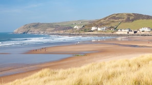A coastal beach with grassy dunes, a sandy shore, and a hill with buildings in the background.