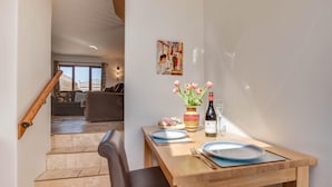 Une salle à manger avec une table en bois dressée pour deux personnes, un vase de tulipes et une bouteille de vin. Un escalier mène à un salon où se trouvent un canapé et un tableau accroché au mur.