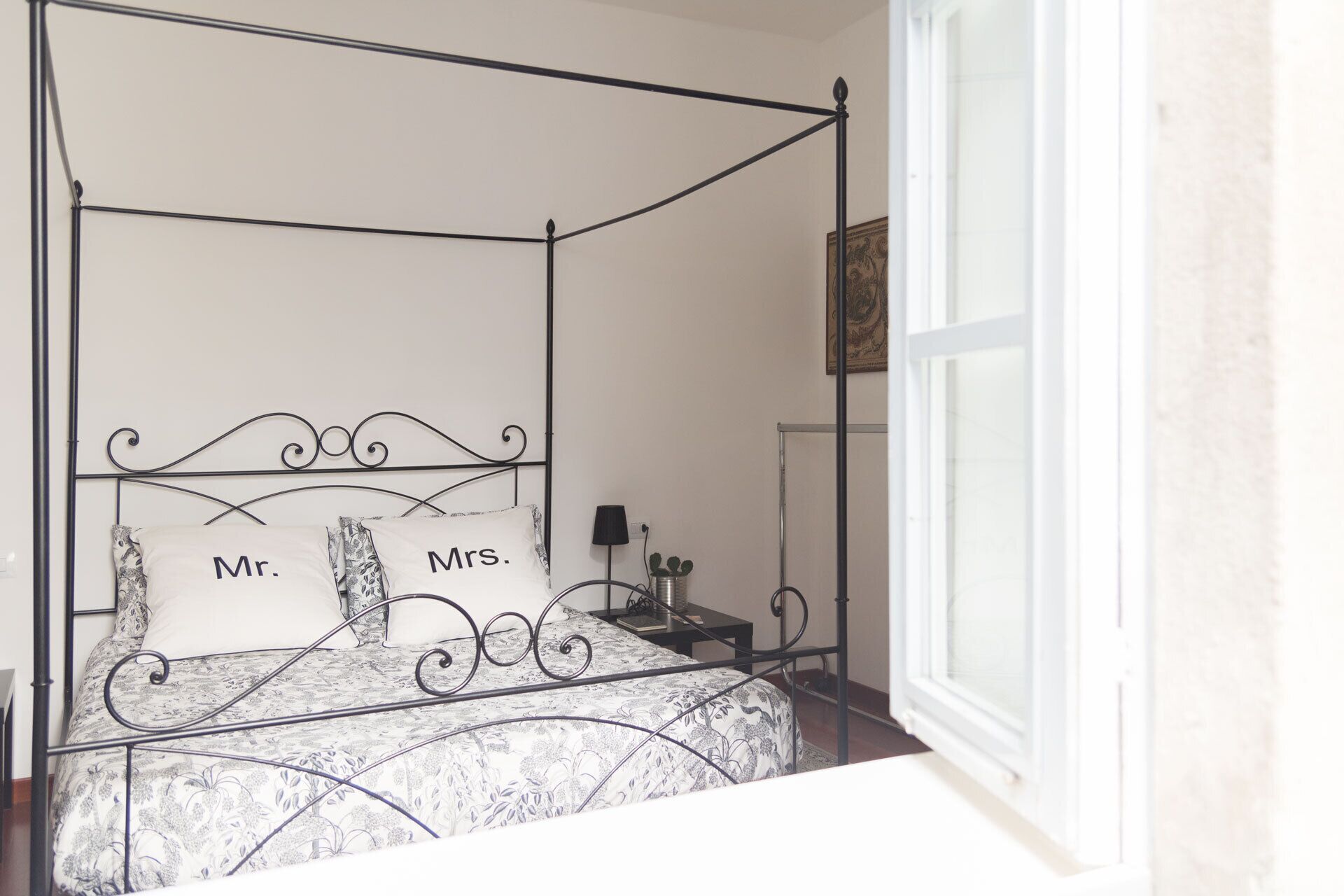 A four-poster bed with "Mr." and "Mrs." pillows, a bedside table with a lamp, and a window with a view.
