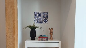 Et hvidt hjørne med en væg med struktur, et lille hvidt bord med en plante og bøger, samt en statue.