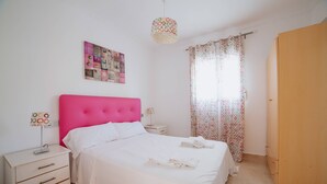 A bedroom with a bed, bedside tables, a nightstand, a hanging lamp, a door, and a window with curtains. - Villa Benicuco 24 - PlusHolidays (Calpe)
