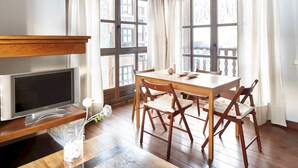 A dining area with a wooden table and chairs, a television on a wooden stand, and a window with curtains.