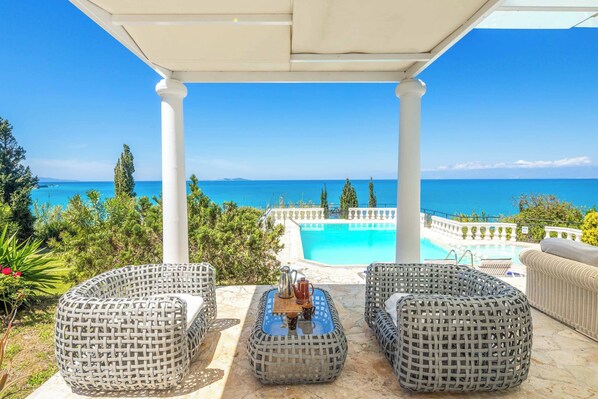Un espacio de estar techado al aire libre con mobiliario de mimbre, piscina y vistas al mar.