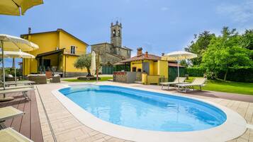 Une piscine avec des chaises longues et des parasols, un bâtiment jaune et, au loin, le clocher d’une église.