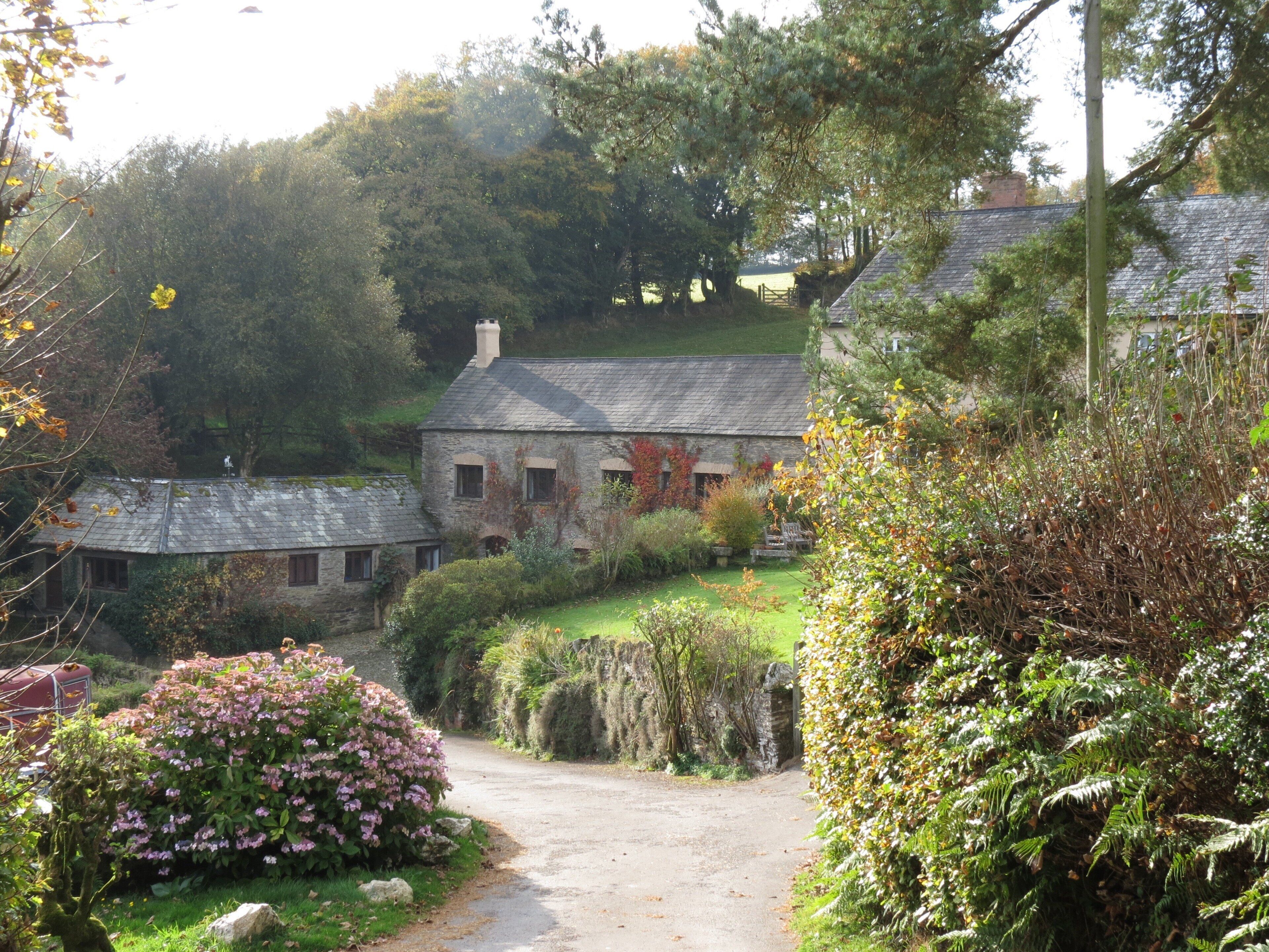 A stone house with a gravel driveway leading to a gate, surrounded by lush greenery and flowering bushes.