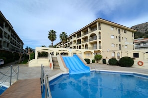 A swimming pool with a blue slide, a multi-story building with balconies, and palm trees.