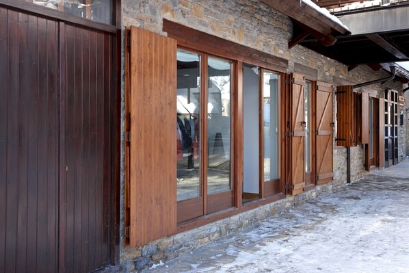 A rustic building with wooden shutters and stone walls, featuring large windows and a snow-covered ground.