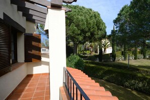 A residential building with a tiled patio, a staircase with a black railing, and a well-maintained garden with trees and shrubs.