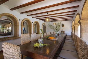 Una larga mesa de comedor con sillas de mimbre, techo de madera con vigas a la vista y vistas a través de ventanas de arco.