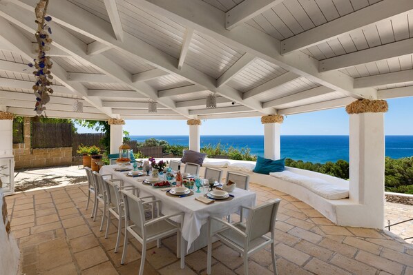 A covered outdoor dining area with a view of the sea, white furniture, and a table set for a meal.