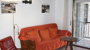 Une pièce avec un canapé rouge, une chaise en bois, une petite table ronde et un lampadaire. Deux photographies en noir et blanc sont accrochées au mur.