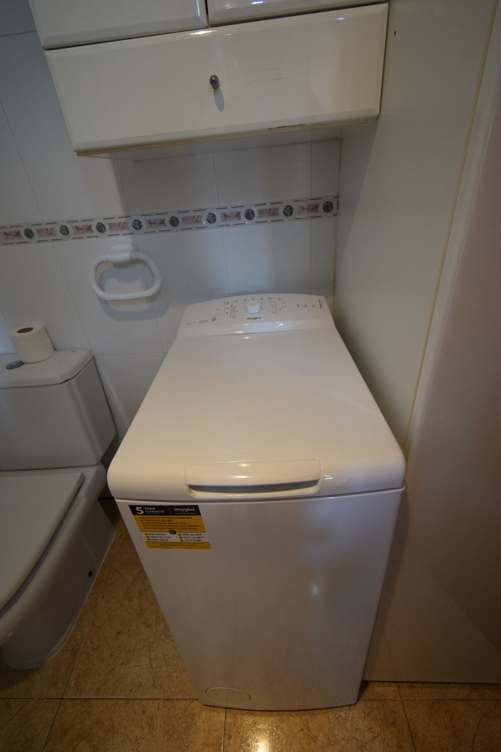 Een compacte, witte wasmachine in een kleine badkamer met een toilet en een handdoekhouder.