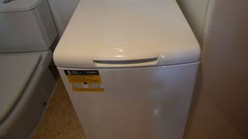 Een compacte, witte wasmachine in een kleine badkamer met een toilet en een handdoekhouder.