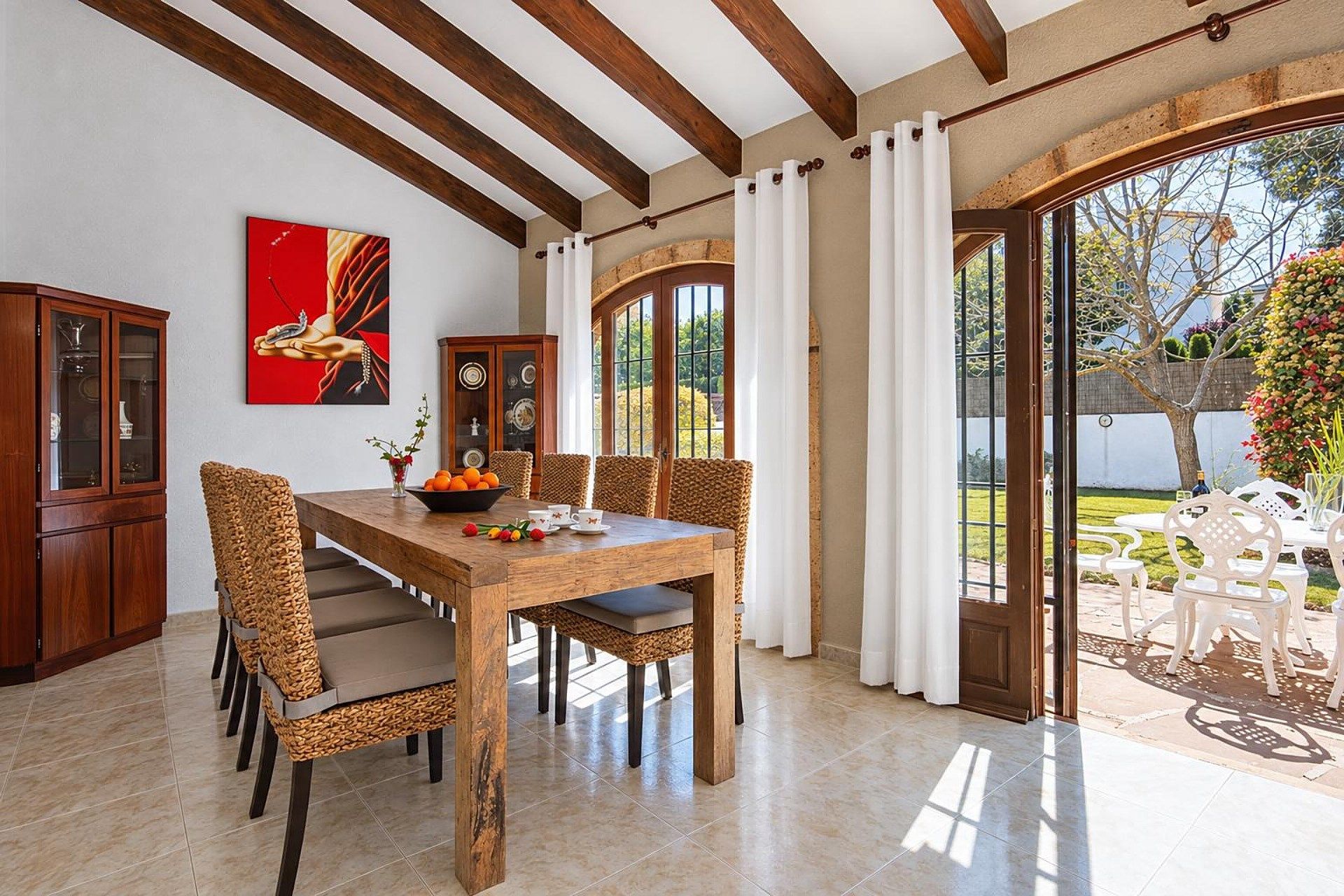 A dining area with a wooden table and chairs, a painting on the wall, and a view through a glass door to an outdoor patio.