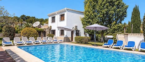 A swimming pool with lounge chairs, a white building with balconies, and a large tree.