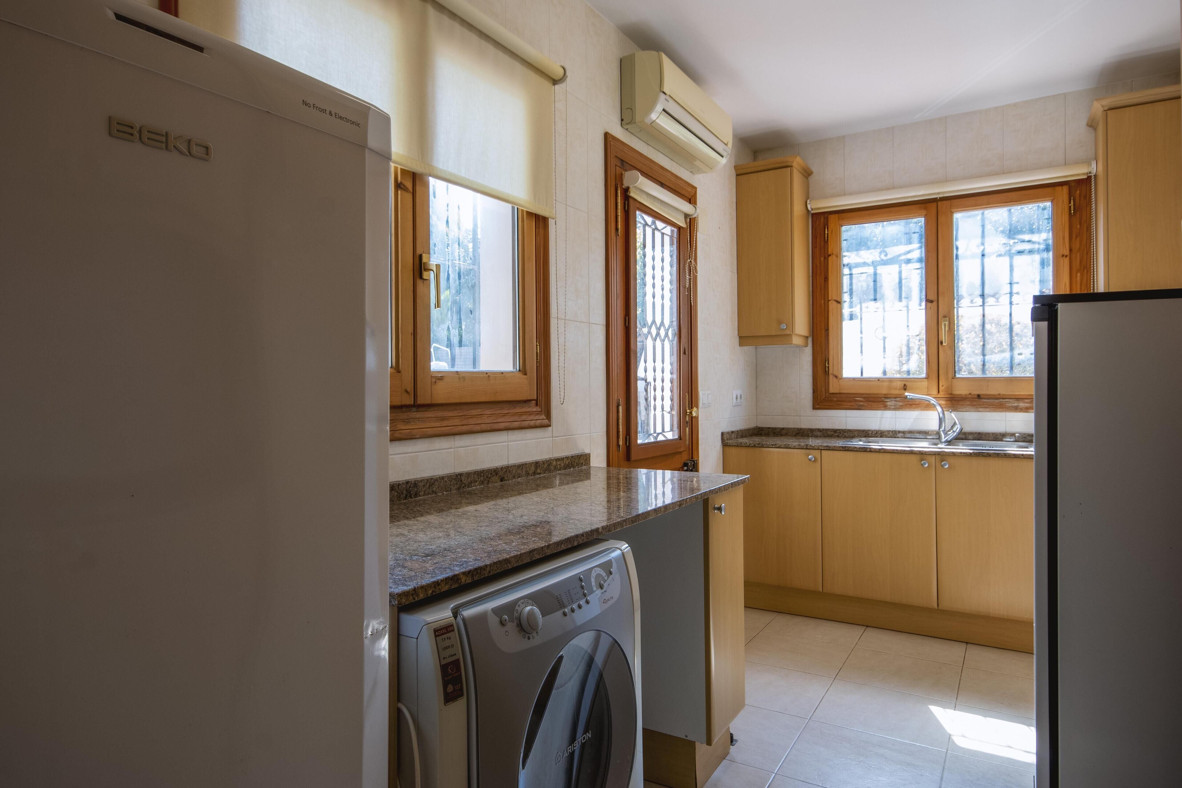Una cocina con lavadora, refrigerador, armarios de madera, una ventana y un equipo de aire acondicionado.