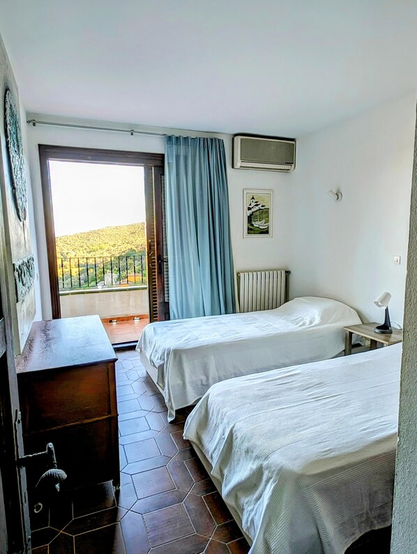 Habitació d'hotel amb dos llits, un taulell de fusta, un radiador, una taula petita amb una làmpada, una finestra amb cortines i un quadre a la paret.