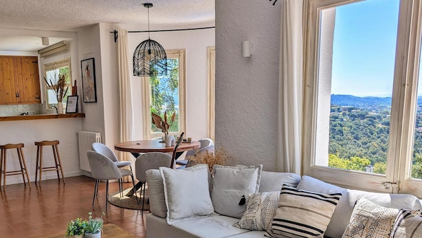 A modern living room with a large window, a sofa, a dining table, and a kitchen area.