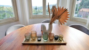 A wooden table with a tray holding various glassware, a white vase with dried plants, and a view of a green landscape through large windows.