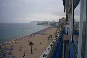 Ein Strand mit Liegestühlen und Sonnenschirmen, eine Promenade mit Palmen und eine Stadtlandschaft im Hintergrund.