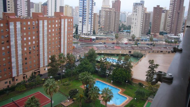 Un edificio de gran altura con piscina en la azotea, rodeado de vegetación y una cancha de tenis.