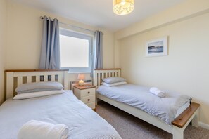 A bedroom with two single beds, a bedside table with a lamp, a window with curtains, and a framed picture on the wall.