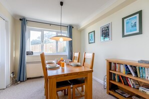 Uma zona de refeições com mesa e cadeiras de madeira, uma estante com livros, uma janela com cortinas e um radiador.