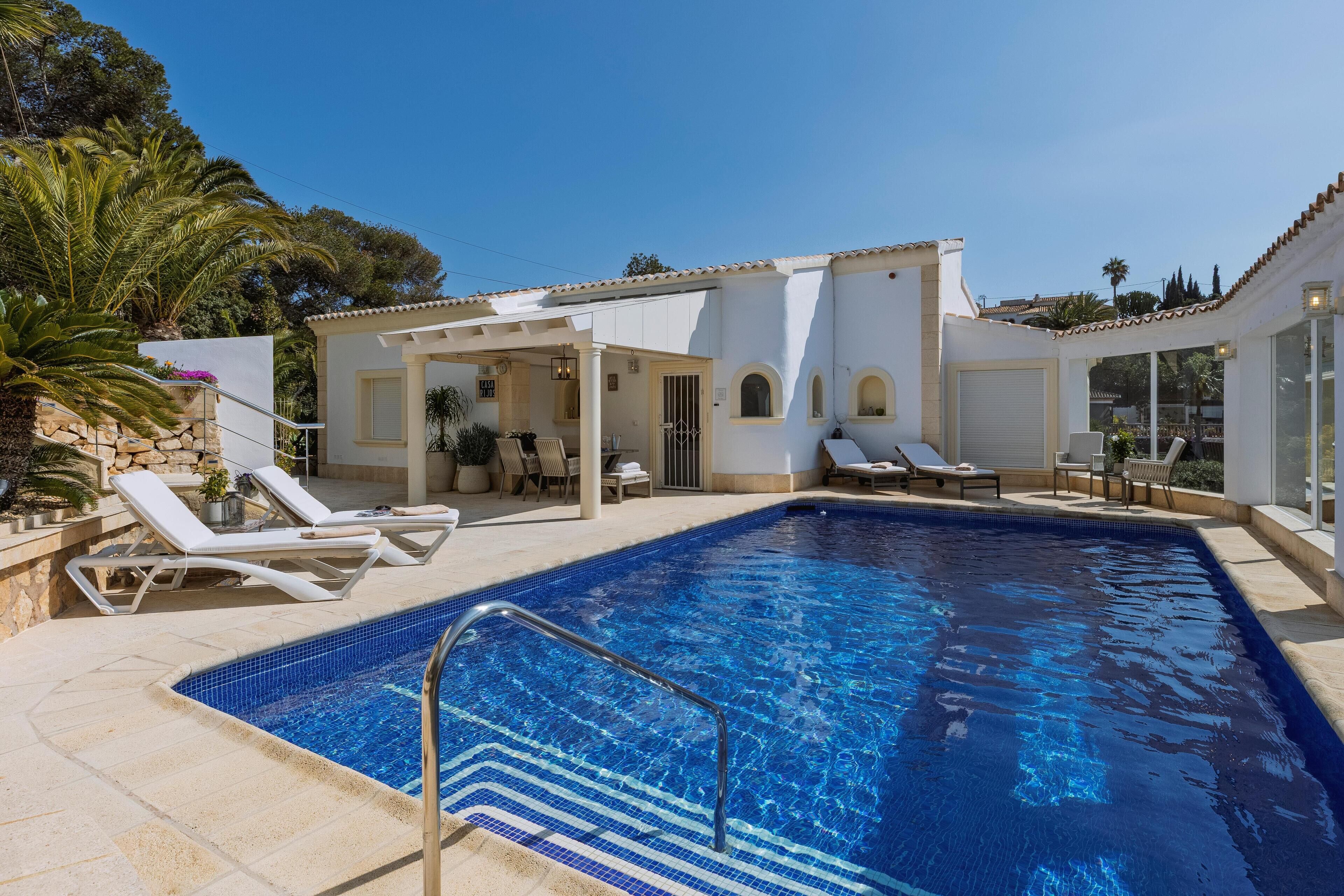 A swimming pool with lounge chairs, a covered patio, and a stone wall.