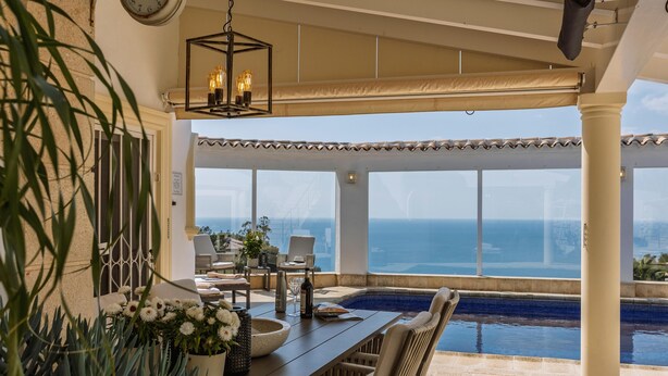 Ein sonnendurchfluteter Essbereich im Freien mit Blick auf das Meer, einen Pool und ein großes Fenster.