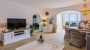 Ein modernes Wohnzimmer mit Flachbildfernseher, weißen Schränken, Korbmöbeln und Blick aufs Meer.