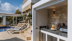 Eine moderne Außenküche mit Steinwand, ein Pool mit Liegestühlen und eine Treppe zu einer überdachten Terrasse.