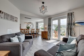 A modern living room with a grey sofa, wooden dining table, and a large window with curtains.