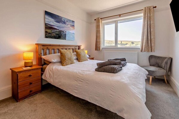 A bedroom with a bed, a nightstand, a lamp, a chair, a window with curtains, and a painting on the wall. - 5 Bed in Woolacombe (oc-heath) (Woolacombe)