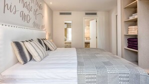 Um quarto com cama, parede com texto motivacional, guarda-roupa com roupas dobradas e um banheiro visível por uma porta aberta.