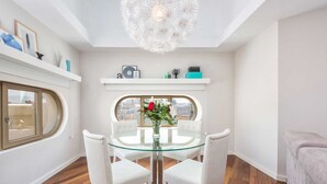 Uma área de refeições com mesa de vidro e cadeiras brancas, um vaso com flores e vista pela janela.