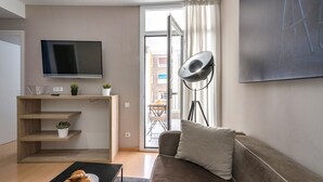 A modern living room with a flat-screen TV, a sofa, a coffee table with croissants, and a balcony with a view of buildings.