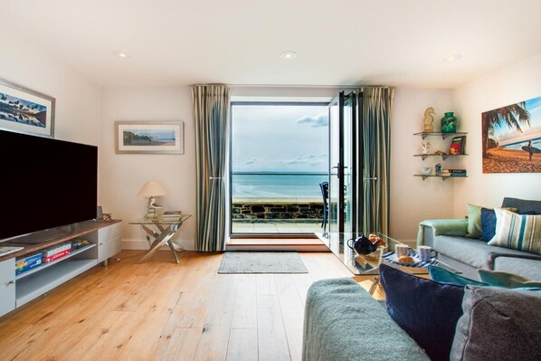 Ein Wohnzimmer mit einem großen Fernseher, einem Glastisch, einer Couch und Blick auf den Strand durch eine Glastür.