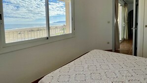 A bedroom with a large window overlooking a beach, a bed with a patterned bedspread, and a door leading to another room. - ROCAMAURA I-A-3-1 (Estartit)