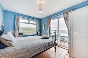 A bedroom with a bed, a bedside table, a window, and a view of the sea. - 4 Bed in Woolacombe (oc-n2116) (Woolacombe)