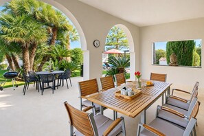 An outdoor patio with a wooden table and chairs, surrounded by palm trees and a Buddha statue.