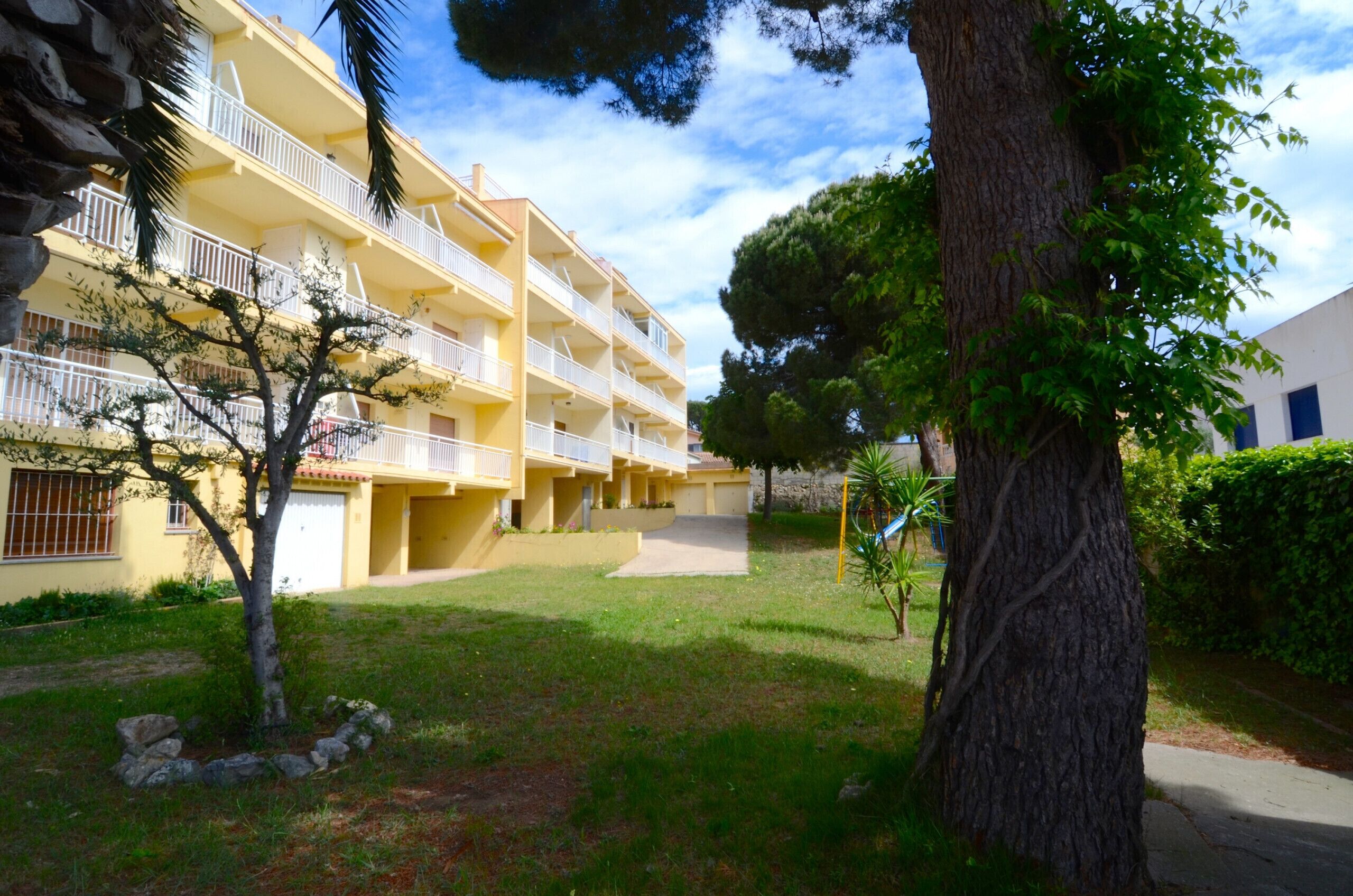 A multi-story yellow building with balconies, surrounded by greenery and trees.